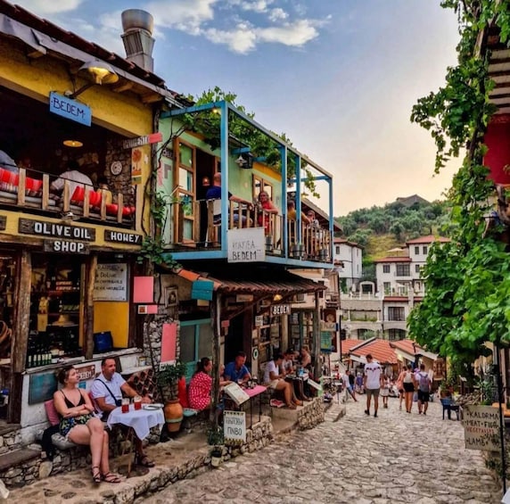 A cobblestone street with shops, including an olive oil shop and a honey shop, and people sitting outside. 1