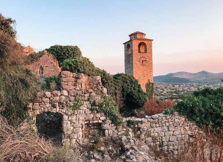 Ancient ruins with a clock tower, surrounded by greenery and overlooking a distant town. 2