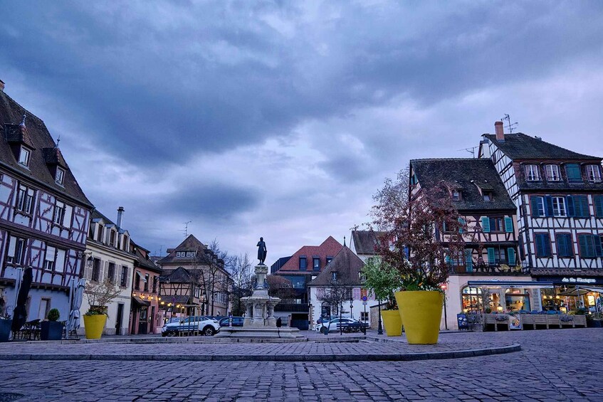 A cobblestone square with traditional half-timbered buildings, a statue, and a large yellow planter. 5