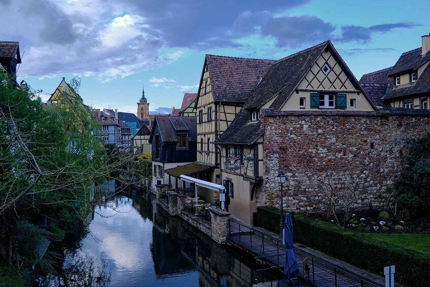 A narrow canal flanked by traditional half-timbered houses with a stone wall on one side and a church steeple in the background. 1