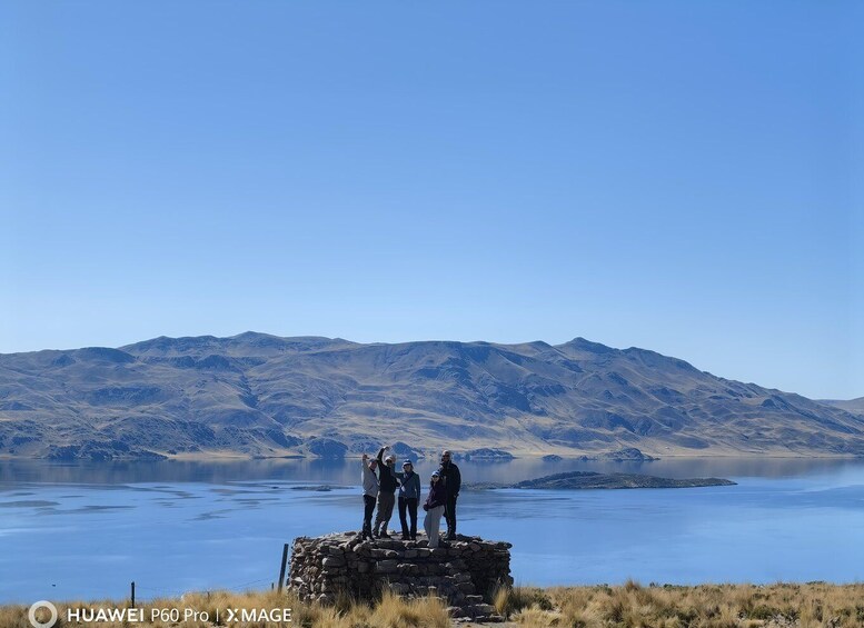 A group of people standing on a stone structure by a calm lake with mountains in the background. 3