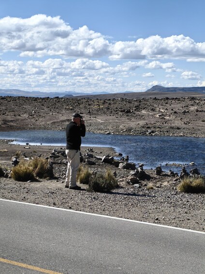 A person taking a photo near a rocky shoreline with a body of water and distant mountains. 3