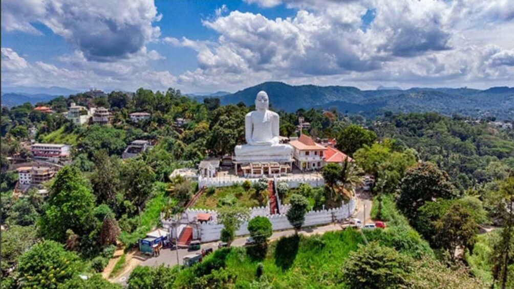 A large white statue on a hill with a temple complex, surrounded by greenery and houses. 1