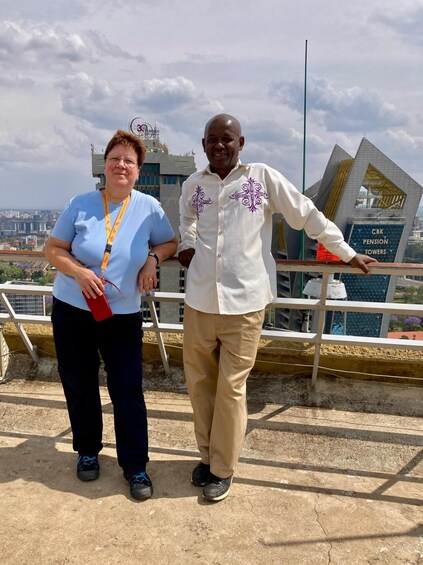 Two people standing on a rooftop with a cityscape and a building labeled 'CBK Pension Towers' in the background. 2