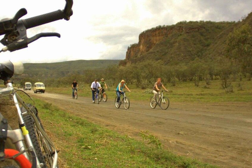 A dirt road with cyclists and a vehicle in the distance, surrounded by greenery and a cliff. 5