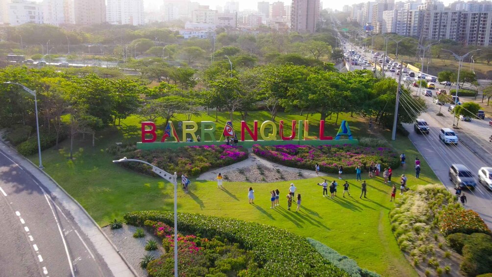 A park with a large sign reading 'BARANQUILLA' surrounded by greenery and a busy road. 1