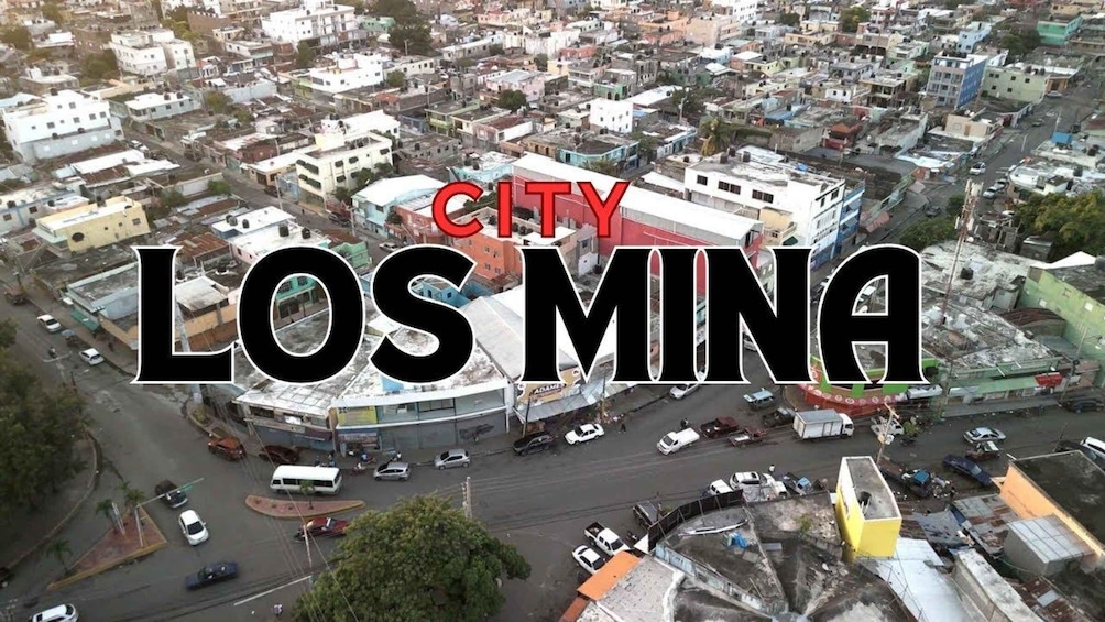 An aerial view of a densely populated urban area with a mix of residential and commercial buildings, a prominent red-roofed structure, and a busy street with vehicles. 1