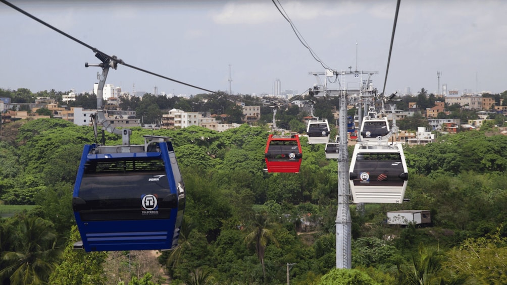 A cable car system with blue, red, and white cars suspended by cables, moving over a lush green landscape with buildings in the background. 5