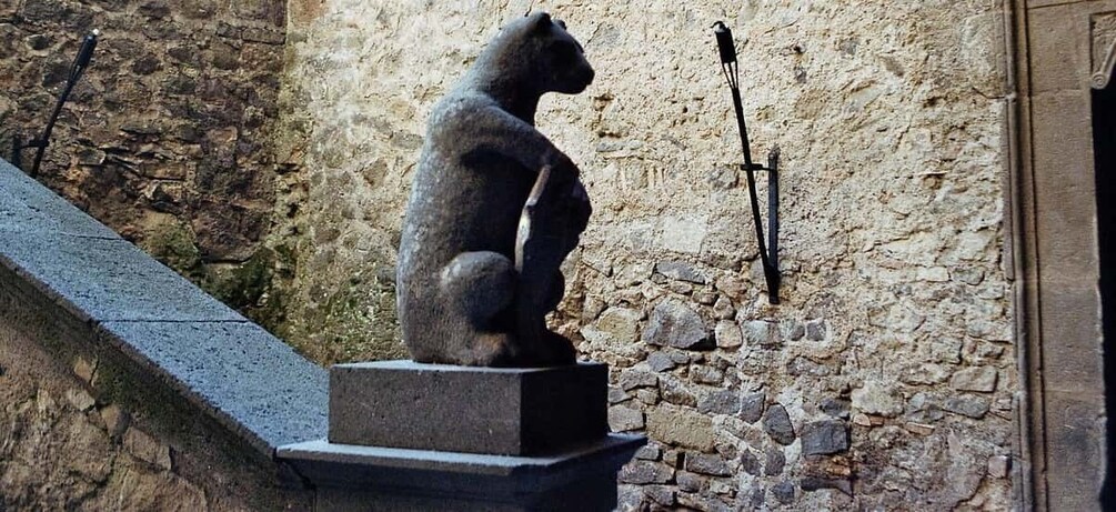 A stone bear statue on a pedestal, set against a stone wall with a door and a black metal pole. 3