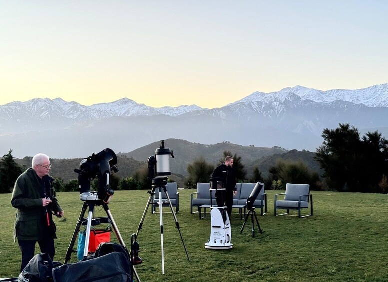 A person with a telescope on a tripod, another person with a telescope, and a chair in a grassy field with mountains in the background. 3