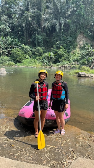 Two people in life jackets and helmets standing by a pink raft on a riverbank with dense greenery in the background. 3