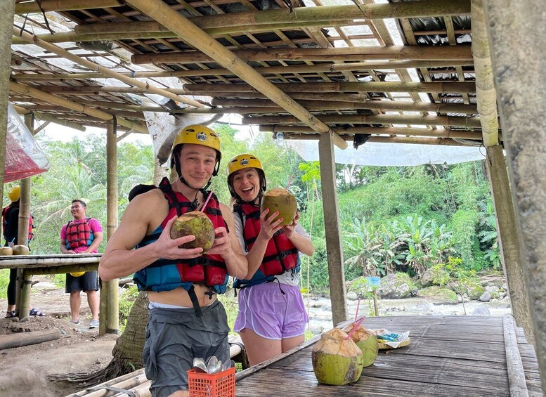 Two people wearing helmets and life vests, holding coconuts, with a wooden structure and greenery in the background. 5