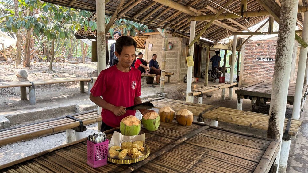 A person cutting coconuts on a bamboo table in an outdoor setting with a brick building in the background. 4
