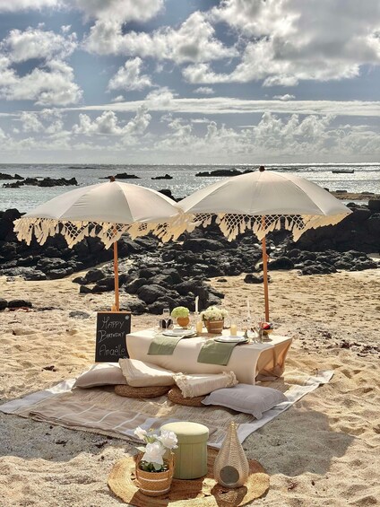 A beach picnic setup with two umbrellas, a table with plates, and a sign that reads 'Happy Birthday Anaelle.' 1