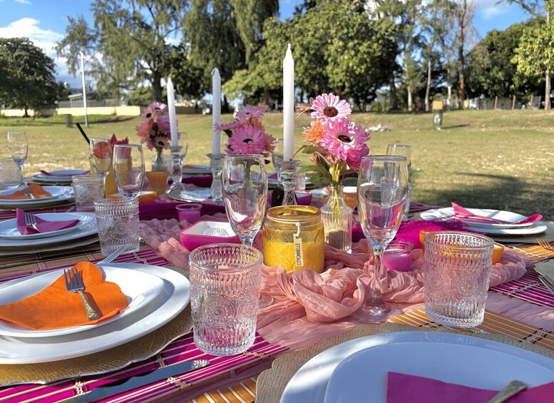 An outdoor picnic with a table set for two, featuring pink and orange tablecloths, clear glassware, and floral decorations. 4