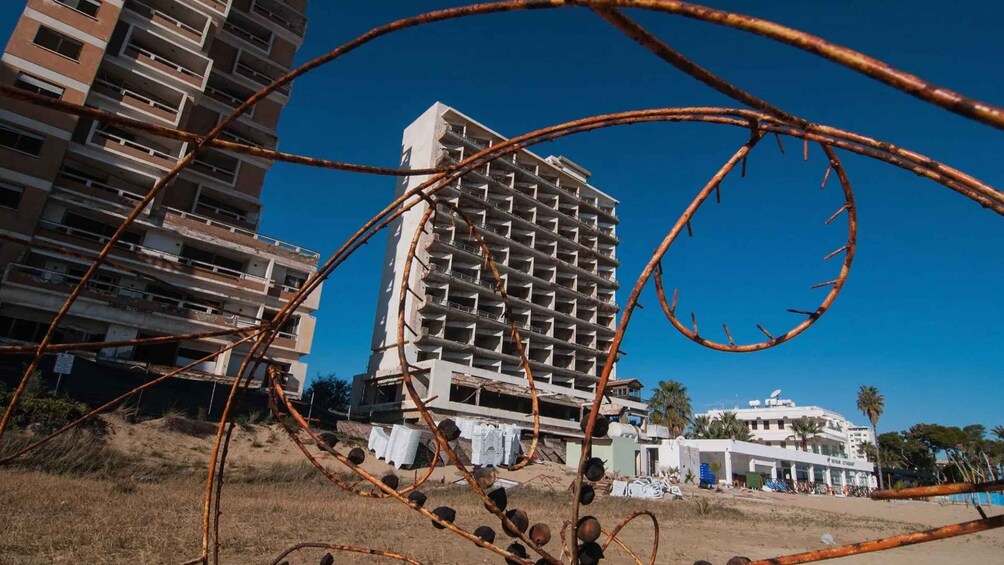 A partially constructed high-rise building with visible scaffolding, surrounded by dry grass and palm trees, under a clear blue sky. 1