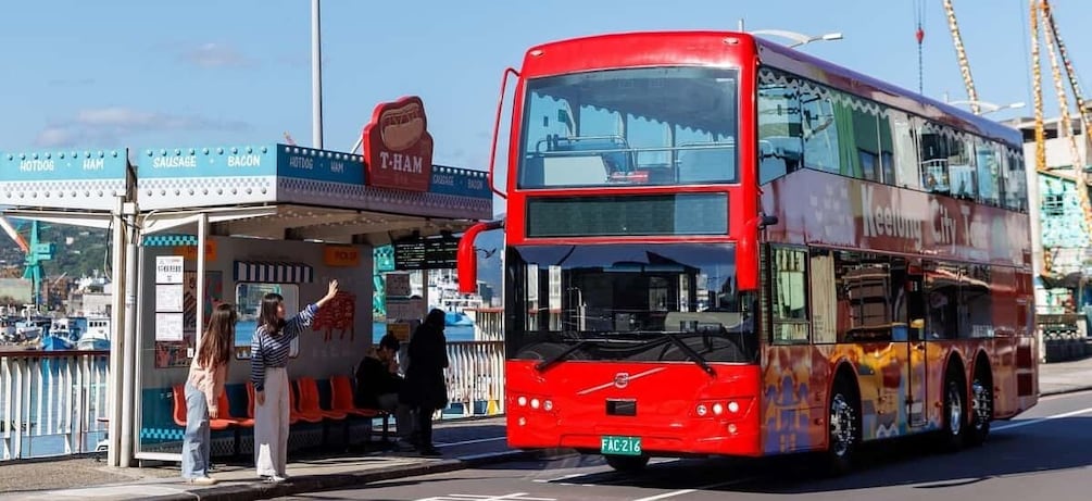 A red double-decker bus parked at a bus stop with a sign for T-Ham and a waterfront in the background. 1