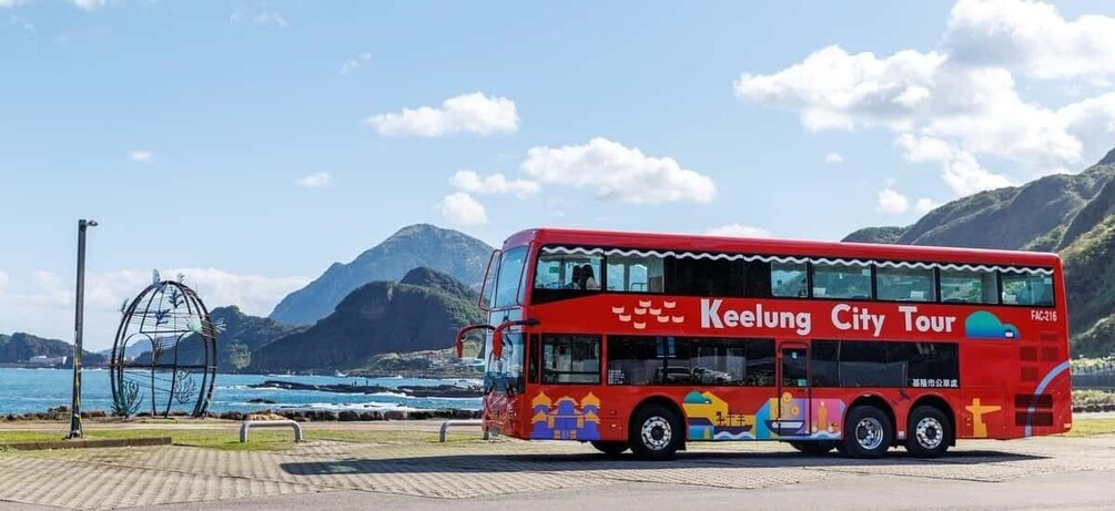 A red double-decker bus with 'Keelung City Tour' written on the side, parked near a coastal area with mountains in the background. 3