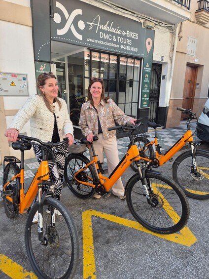 Two people standing next to orange e-bikes in front of a building with signage for e-bikes and tours. 1
