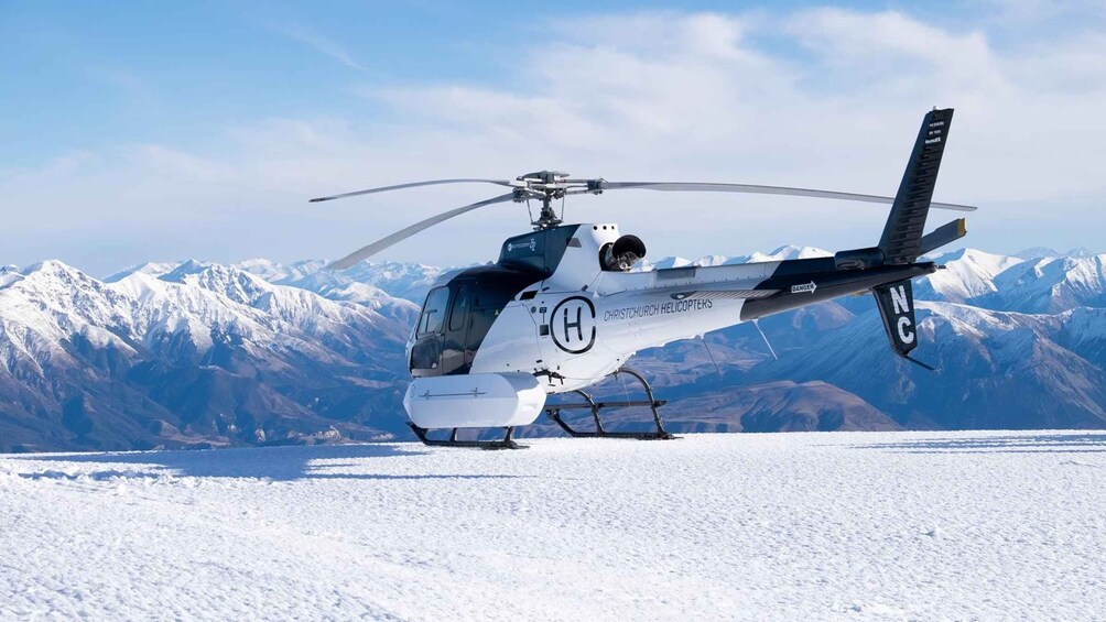 A helicopter with 'CHRISTCHURCH HELICOPTERS' on its side, parked on a snow-covered field with mountains in the background. 1