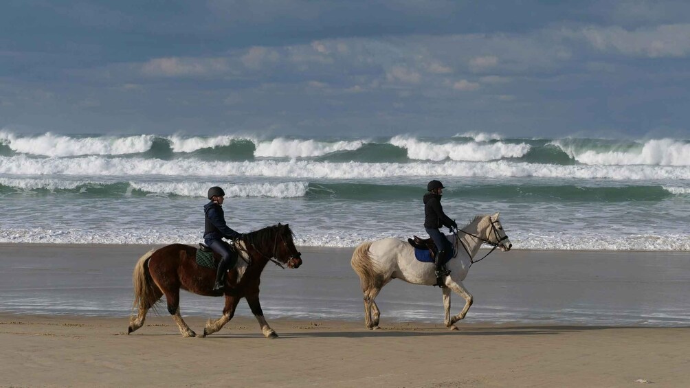 Two riders on horses along a beach with waves in the background. 2