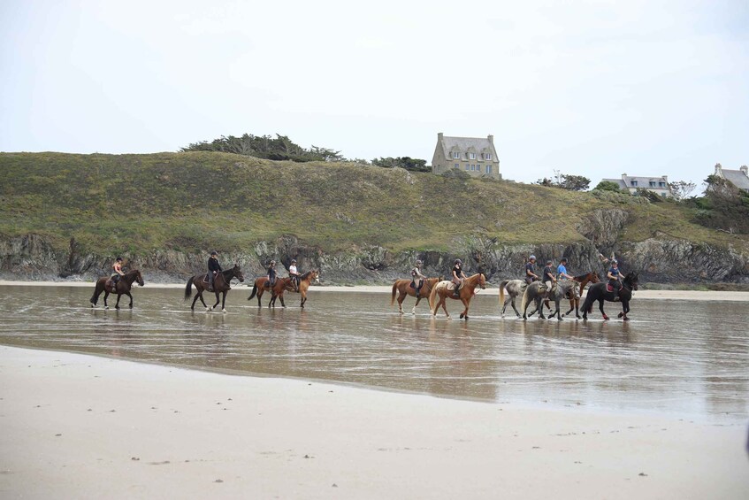 A group of people riding horses along a beach with a hill and a house in the background. 3