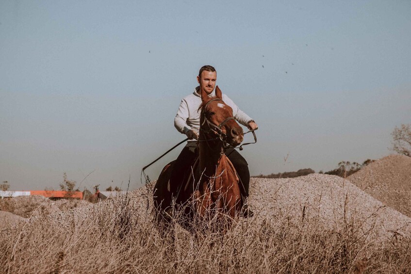 A person riding a horse in a field with dry grass and a pile of sandbags in the background. 1