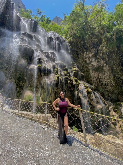 A person standing by a cascading waterfall with a rocky cliff and greenery in the background. 4