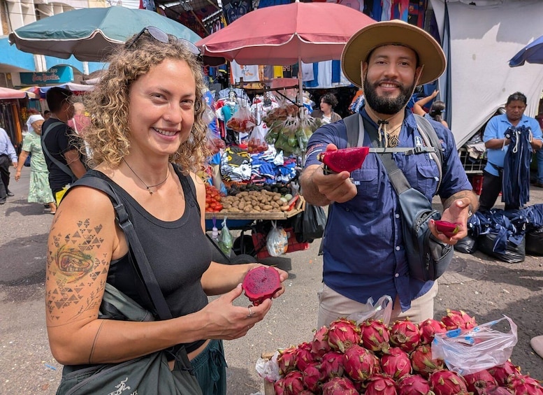 A person holding a dragon fruit and another person with a hat, standing in a market with various goods and a pile of dragon fruits on the ground. 1