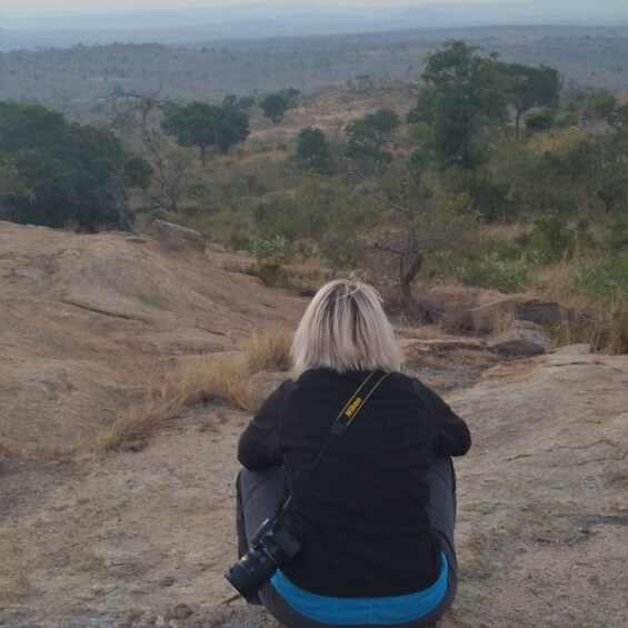 A person sitting on a rock with a camera, overlooking a dry landscape with sparse trees. 1