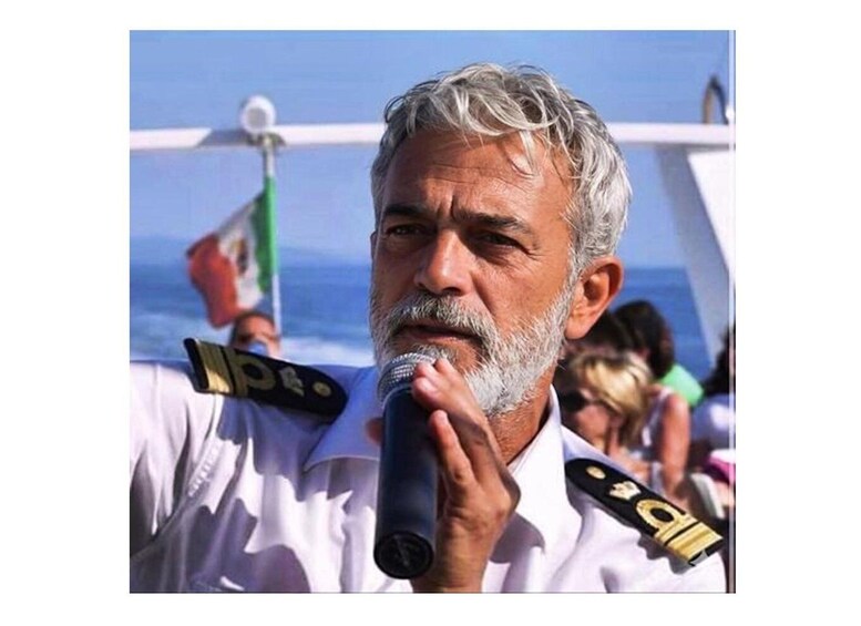 A person in a white uniform with a microphone, standing on a boat with a flag in the background. 1