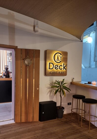 A cafe-bar interior with a wooden door, a sign reading 'Deck Cafe-Bar,' a potted plant, and two bar stools. 2