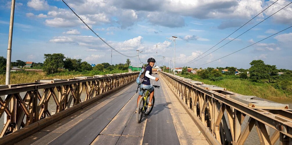 A person riding a bicycle on a wooden bridge with metal railings, surrounded by greenery and houses. 2