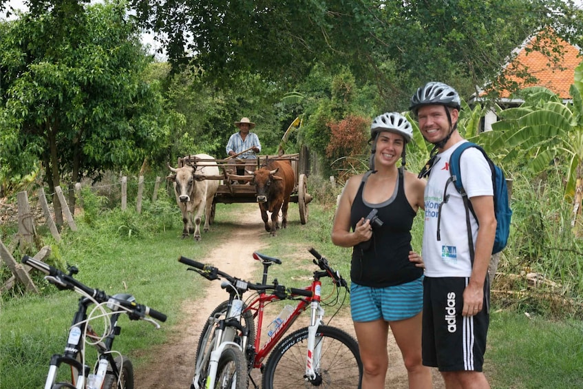 Two people standing by a dirt path with bicycles, a man driving a cart pulled by oxen, and lush greenery in the background. 4