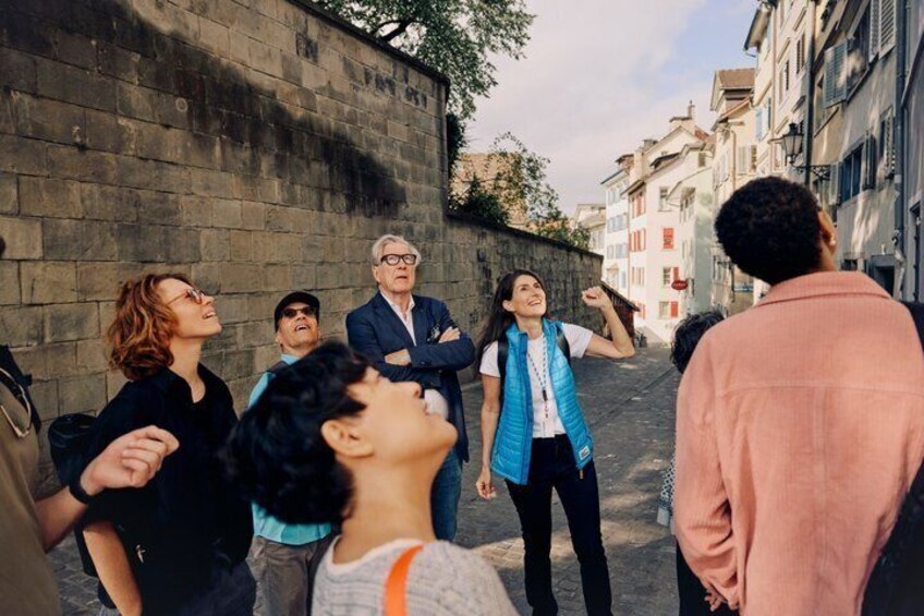 A group of people standing on a street with a stone wall and buildings in the background. 2