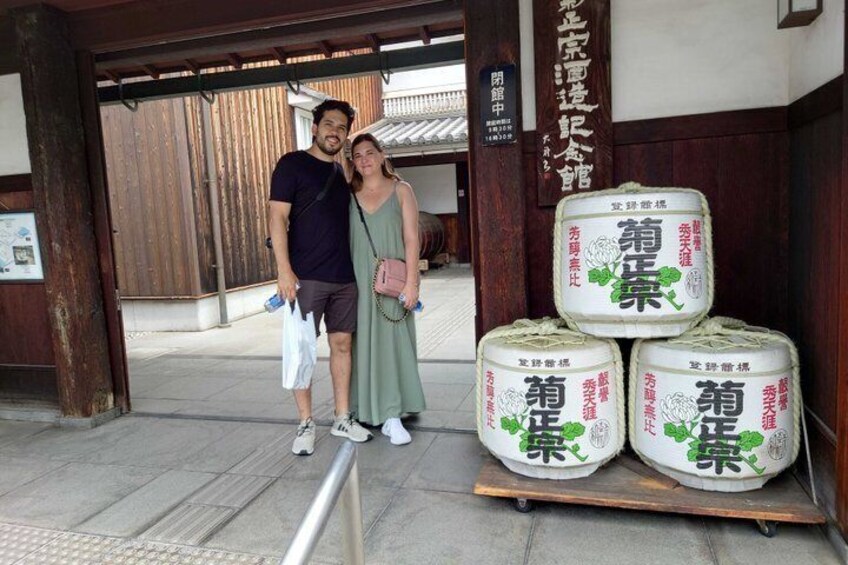 A man and a woman standing in front of a traditional Japanese building with wooden pillars and a tiled roof. There are large sacks with Japanese text and a wooden cart with more sacks. 2