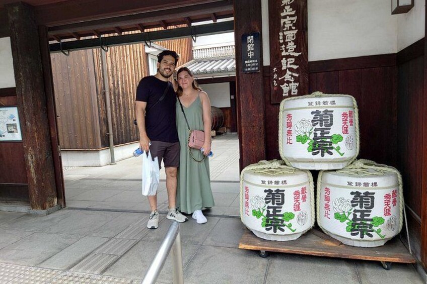 A couple standing in front of a traditional building with wooden doors and a signboard. There are large sacks with Japanese characters on a wooden pallet. 2