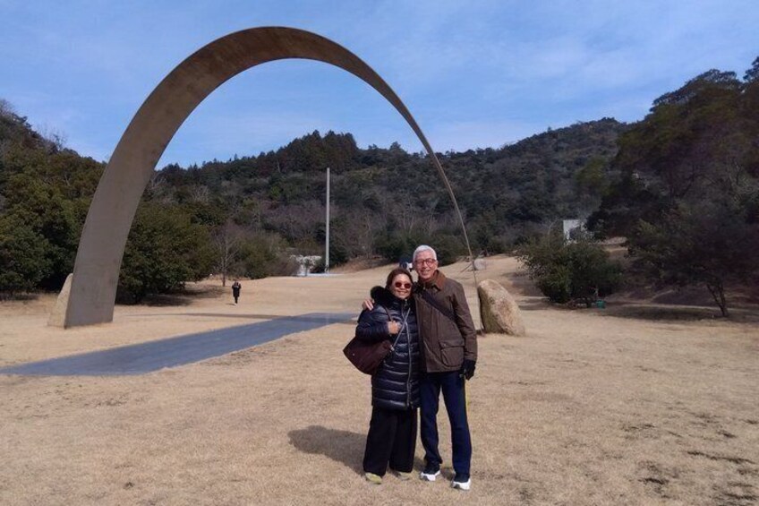 A couple standing in front of a large arch structure in a park with trees and hills in the background. 1