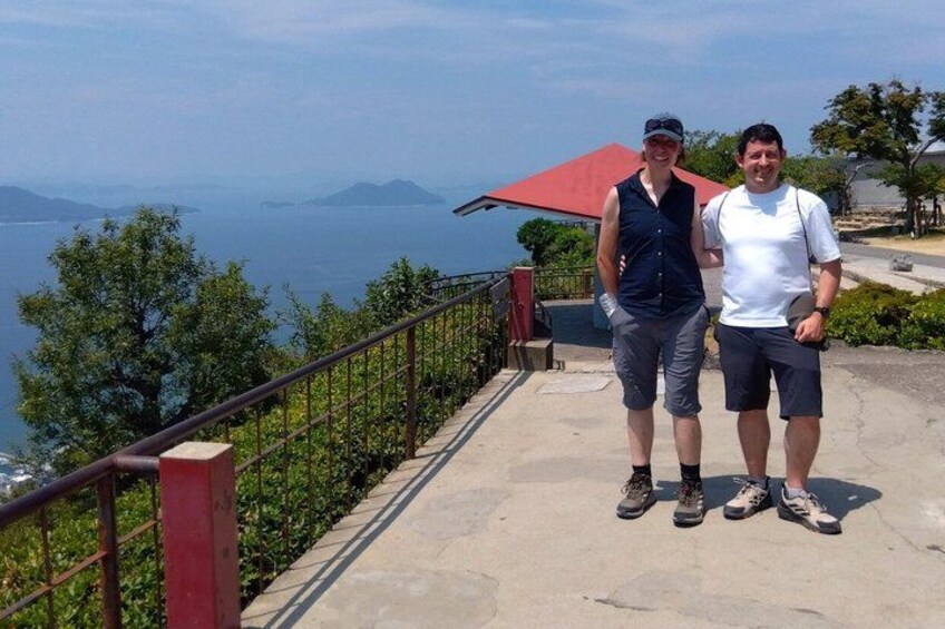 Two people standing on a concrete path with a railing, overlooking a body of water and distant mountains. 2