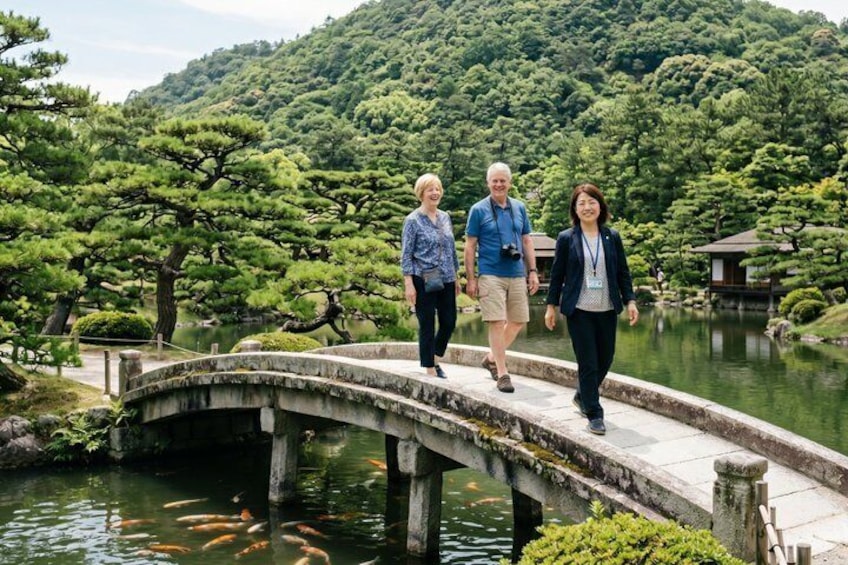 A stone bridge over a pond with koi fish, surrounded by lush greenery and a mountainous backdrop. 1