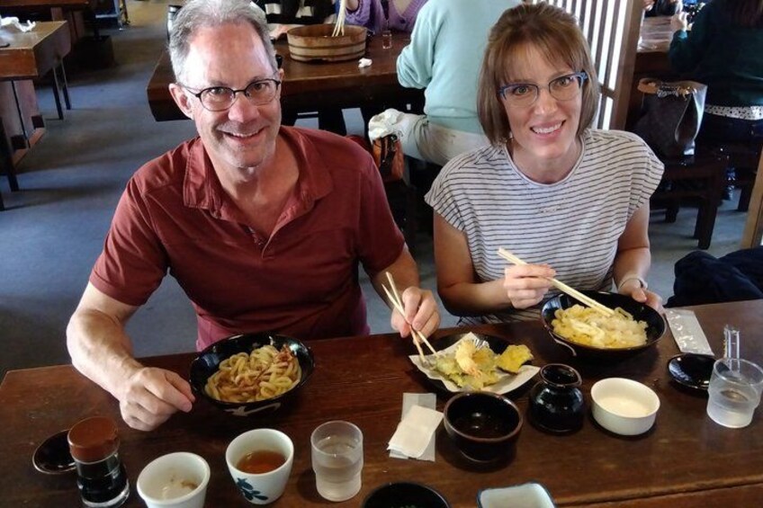 Two people sitting at a table with bowls of food, chopsticks, and small cups. 4