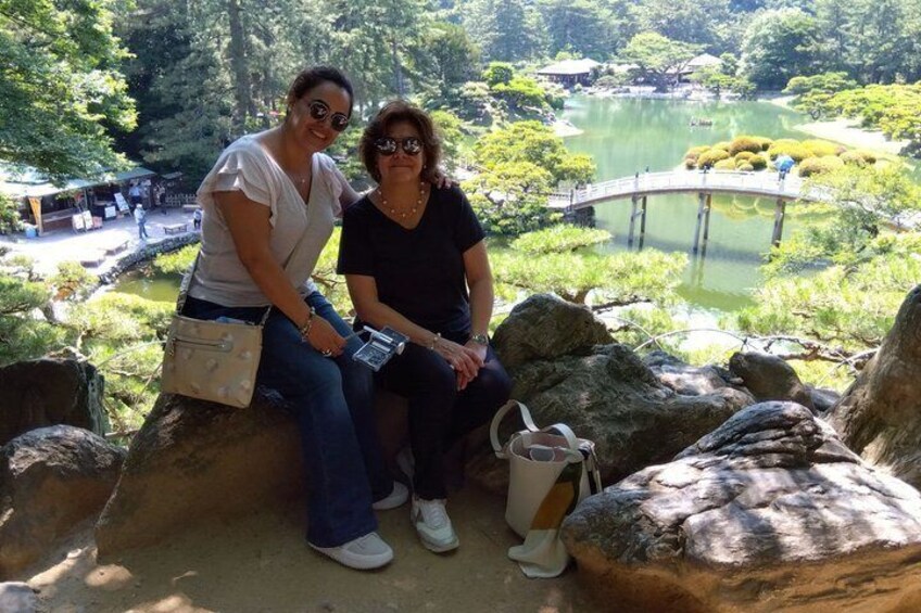 Two people sitting on rocks overlooking a pond with a bridge and Japanese-style landscaping. 5