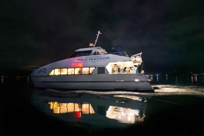 A large white boat with 'TASMANIAN WILD SEAFOOD ADVENTURES' written on its side, docked at night with lights reflecting on the water. 1
