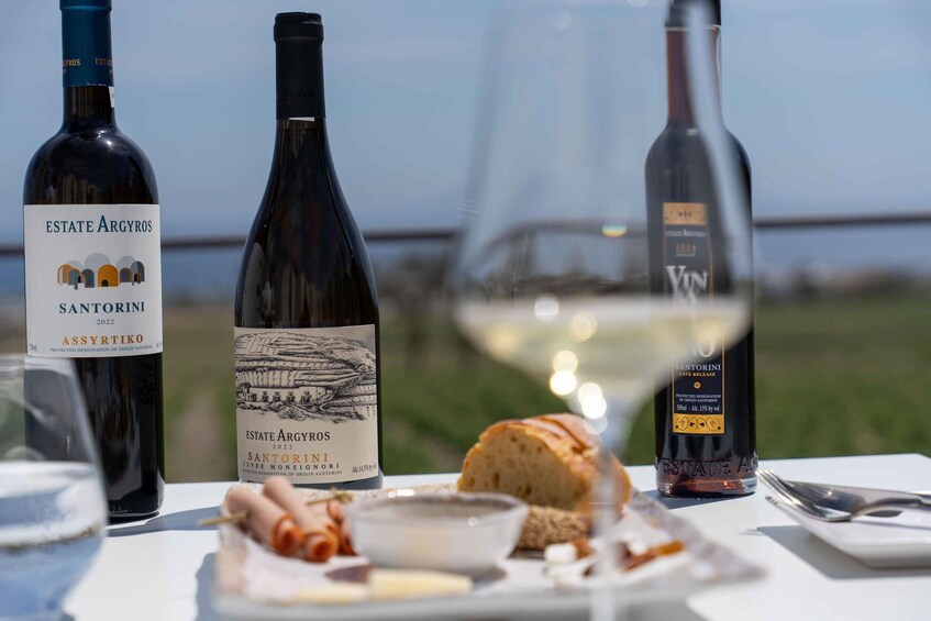 Three bottles of wine, a glass of wine, bread, and a bowl of olives on a table with a coastal view in the background. 3