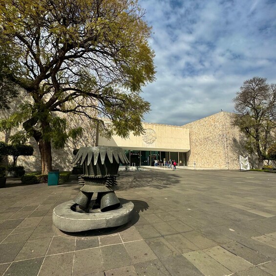 A courtyard with a stone sculpture, trees, and a building with a stone facade. 4
