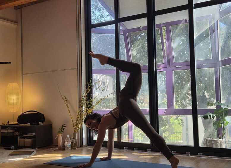 A person performing a yoga pose on a mat in a room with large windows and indoor plants. 3