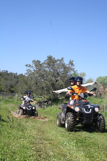 Two people on an ATV in a grassy field with trees in the background. 1