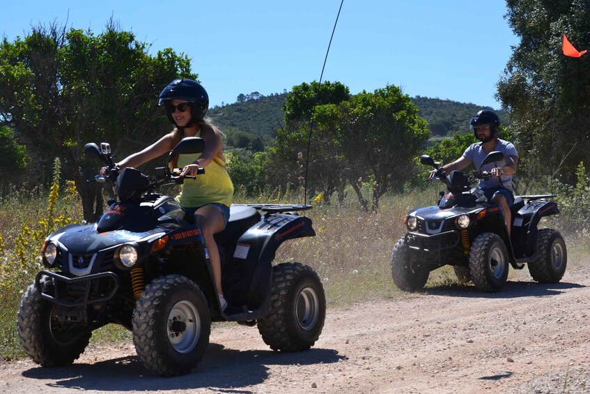 Two people riding ATVs on a dirt road with trees and hills in the background. 3