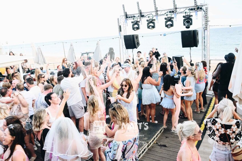 A crowded outdoor event with people on a dock, a stage with speakers, and a body of water in the background. 3