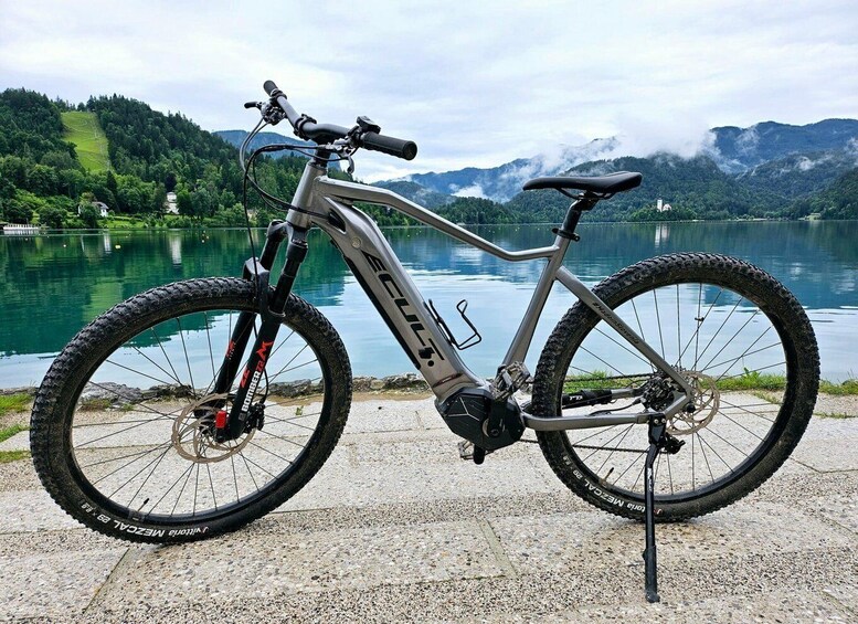 A mountain bike with a water bottle, a rear rack, and a saddle, parked by a lake with mountains in the background. 3